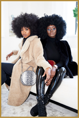 Two adults with large curly hair sit closely on a white couch wearing stylish coats, one in a beige teddy coat with a patterned handbag and the other in a black fur coat with shiny black pants and glasses. Description generated by AI