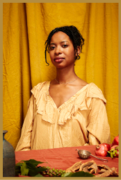 A person with curly hair tied back is wearing a loose, ruffled yellow blouse and sitting at a table with various fruits and a small metal cup in front of a mustard yellow curtain. Description generated by AI