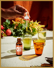 A hand is holding a dropper above a glass filled with green liquid, surrounded by two other glasses, a brown bottle labeled “Oma Botanicals," and leafy greens on a table. Description generated by AI