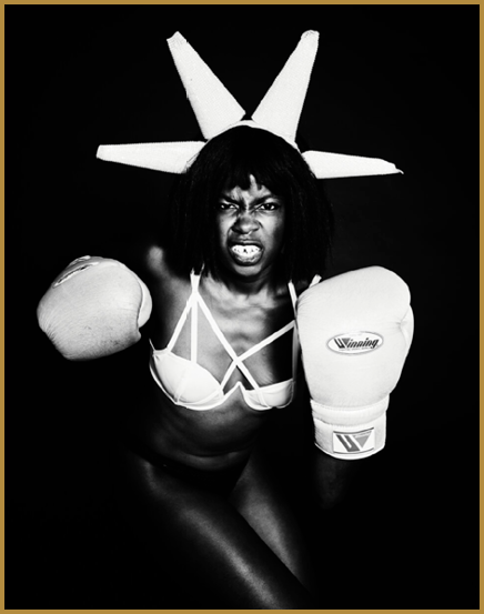 A person wearing boxing gloves and a strappy bikini top poses aggressively with a fierce expression and a headpiece resembling the Statue of Liberty's crown against a black background. Description generated by AI