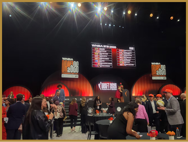 People dressed in business and casual attire gather and converse in a large event space with bright stage lights and screens displaying the WNBA Draft 2023. Description generated by AI