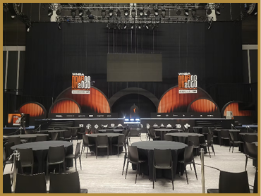 A large stage with a black backdrop and orange semi-circular designs is set up in a spacious venue with round tables and chairs arranged in front of it. Description generated by AI