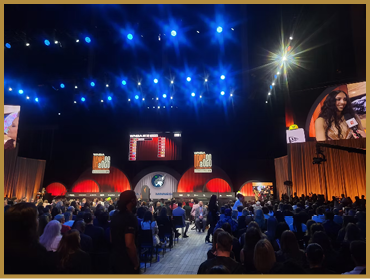 A large audience is seated in a dimly lit auditorium facing a stage with bright blue overhead lights, multiple screens, and a person speaking into a microphone shown on one of the screens. Description generated by AI