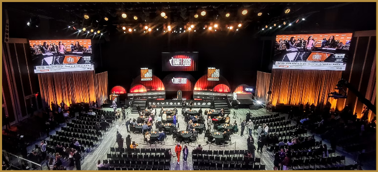 A large indoor event space with rows of chairs facing a stage with multiple screens displaying a panel discussion and people seated and standing around the room. Description generated by AI