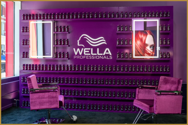 A vibrant purple salon interior features two purple styling chairs facing each other with shelves of hair product bottles and a Wella Professionals logo on the wall between two framed images, one showing a person with bright red hair. Description generated by AI