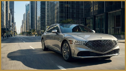 A sleek silver luxury car is driving on a city street lined with tall modern buildings under clear daylight. Description generated by AI